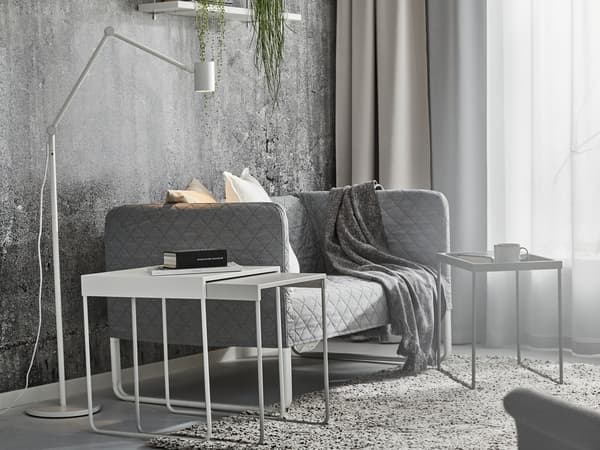 Három szürke és fehér dohányzóasztal áll egy szürke, kétszemélyes KNOPPARP kanapé mellett, a kanapén steppelt takaró és fehér párnák vannak.