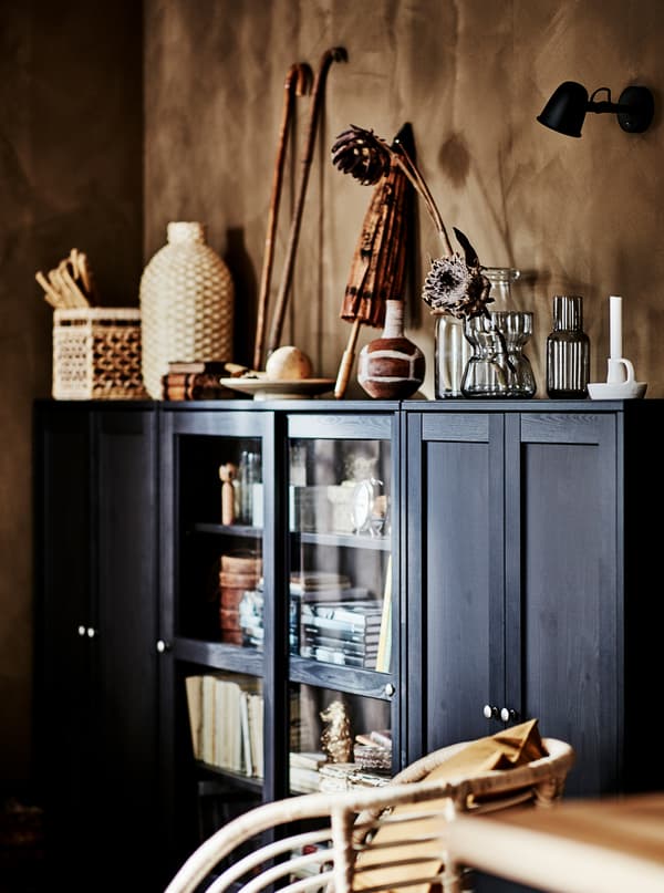 มุมหนึ่งของห้องนั่งเล่น มีตู้ HAVSTA/ฮาฟสต้า ขนาดกว้างสีเข้มที่มีงานศิลปะจากวัสดุธรรมชาติเก็บอยู่ข้างในและวางอยู่ข้างบนตู้