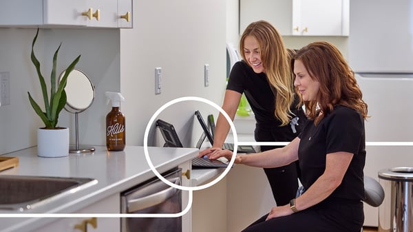 Helle, modern gestaltete Arbeitsküche mit IKEA Möbeln und Dekorationen, zwei Frauen im Gespräch vor einem Tablet, frische grüne Pflanze und braune Sprühflasche als stilvolle Akzente auf der weißen Arbeitsplatte.