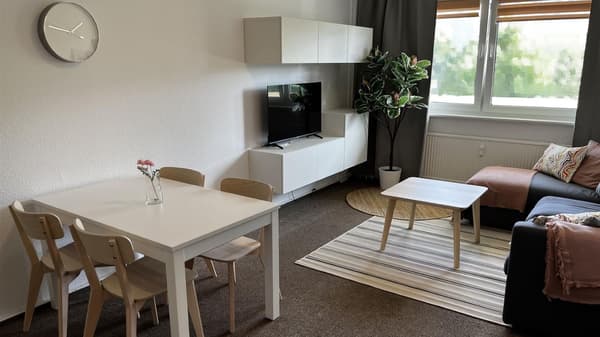 Helles Wohnzimmer mit weißem Tisch und vier Holzstühlen links sowie einer Wanduhr; gegenüber stehen ein TV auf Lowboard, weiße Hängeschränke und eine große Topfpflanze. Rechts befinden sich ein dunkles Sofa mit Kissen und ein kleiner Couchtisch auf einem gestreiften Teppich vor einem Fenster mit grauen Vorhängen und Rollläden.
