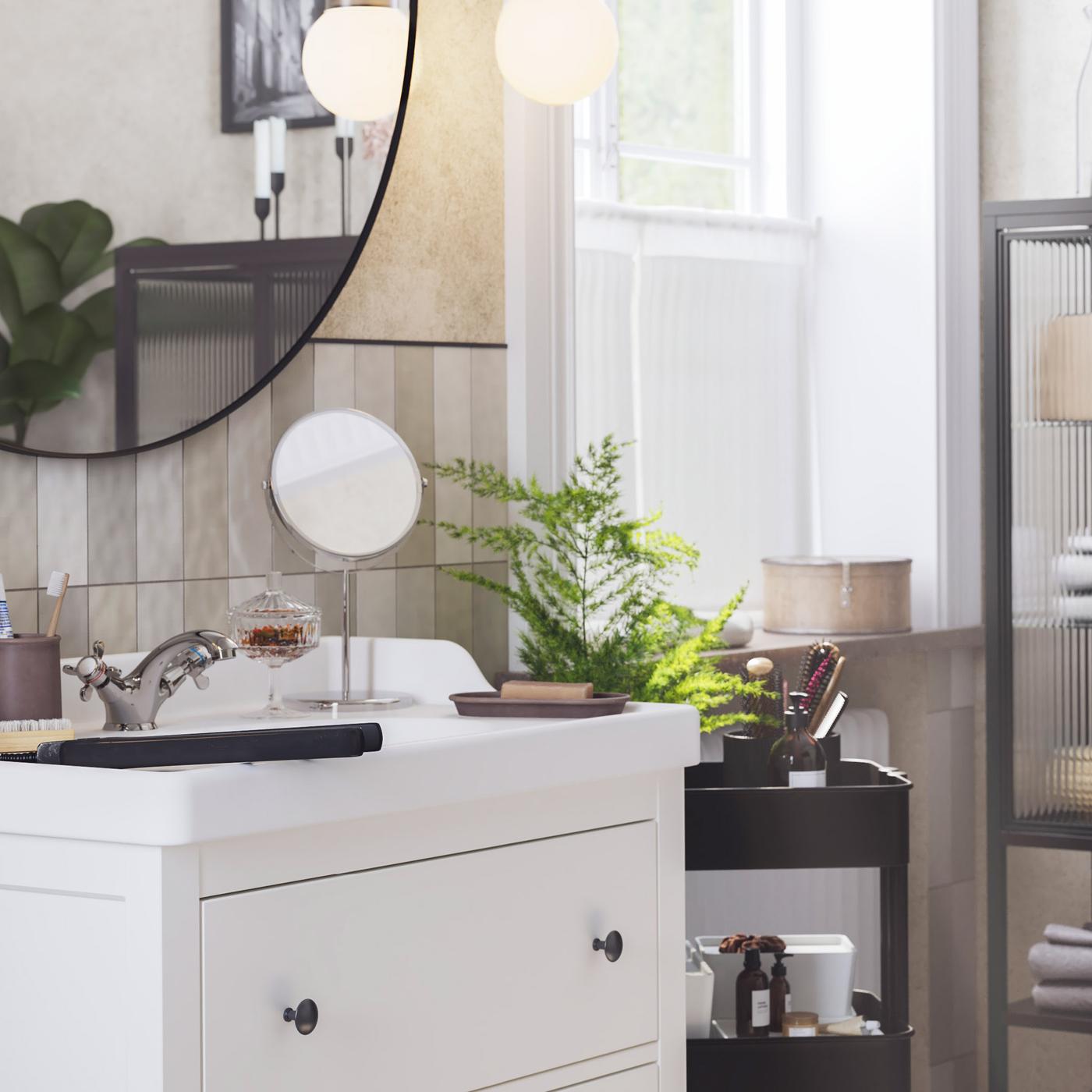 ห้องน้ำที่สว่างและมีหน้าต่าง มีตู้อ่างล้างหน้า HEMNES/เฮมเนส สีขาวและกระจกเงาทรงกลมด้านบน และรถเข็นสีดำอยู่ถัดไป