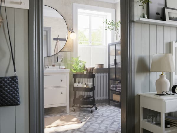 חדר אמבטיה אפור עם חלון גדול ובו מעמד לכיור HEMNES, עגלת מטבח ואחסון RÅSKOG בצבע שחור, ארון עם דלת זכוכית MOSSJÖN.