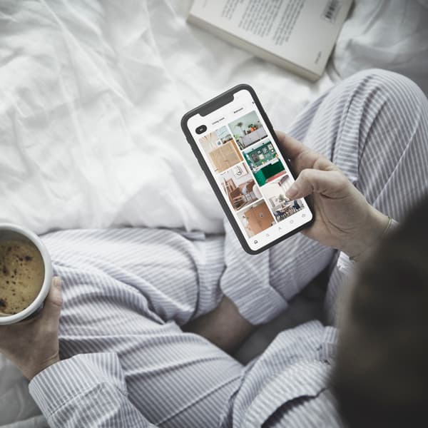 Henkilö istuu sängyllä puhelin ja kahvikuppi kädessään. Puhelimessa on avoimena IKEA-sovellus.