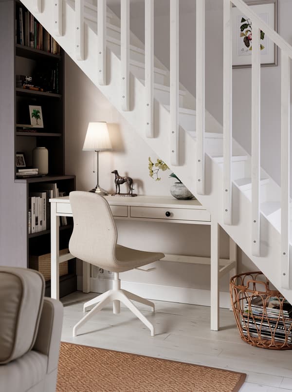 Hier befindet sich ein Arbeitsplatz unter einer Treppe. Er besteht aus einem HEMNES Schreibtisch, einem LÅNGFJÄLL Konferenzstuhl, einer Leuchte und einem hohen Bücherregal.