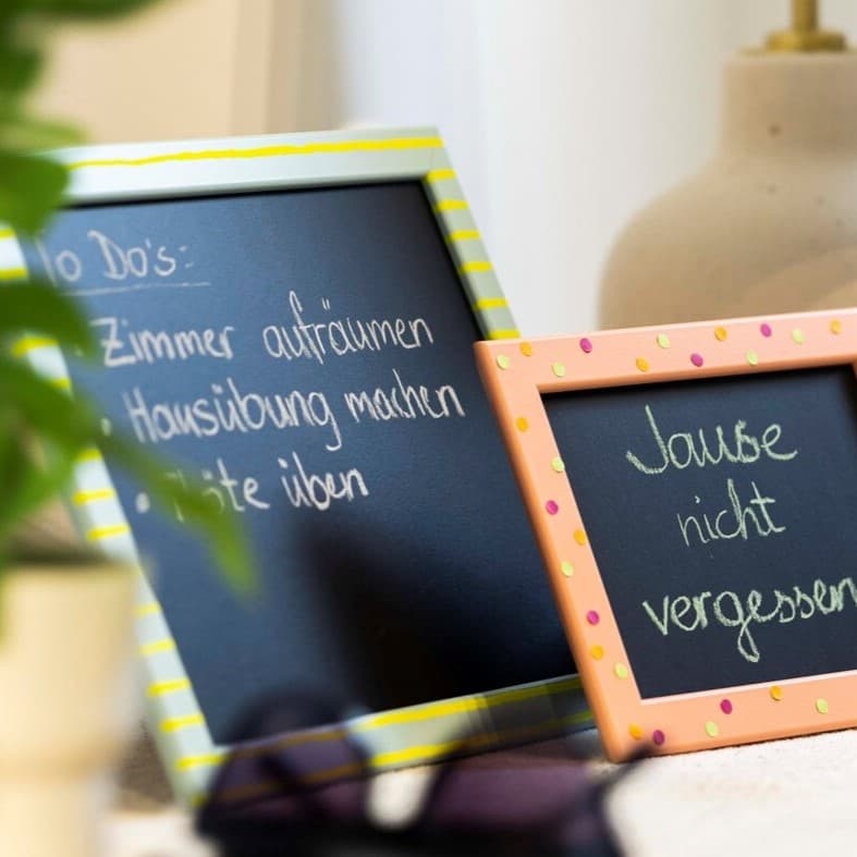 Hier sieht man 2 kleine Tafeln auf einem Tisch stehen mit einer bunten Schrift darauf.