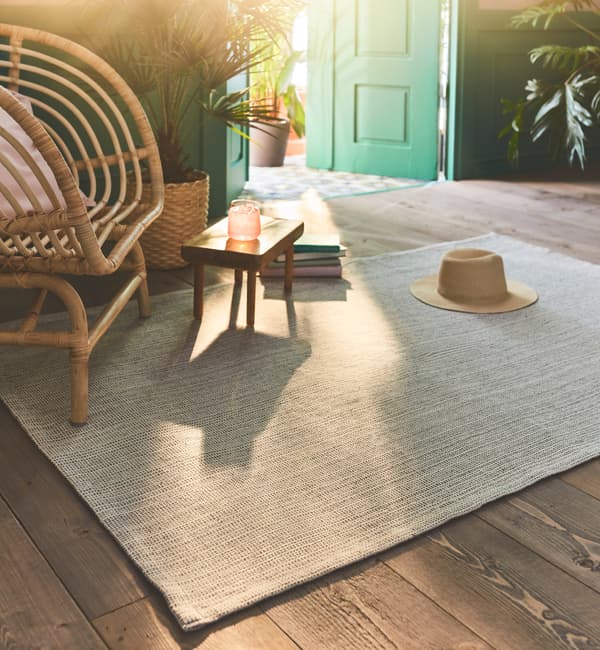 Hladko tkaný koberec TIPHEDE v prírodnej/krémovej farbe na drevenej podlahe vedľa prúteného kresla v slnečnej miestnosti.