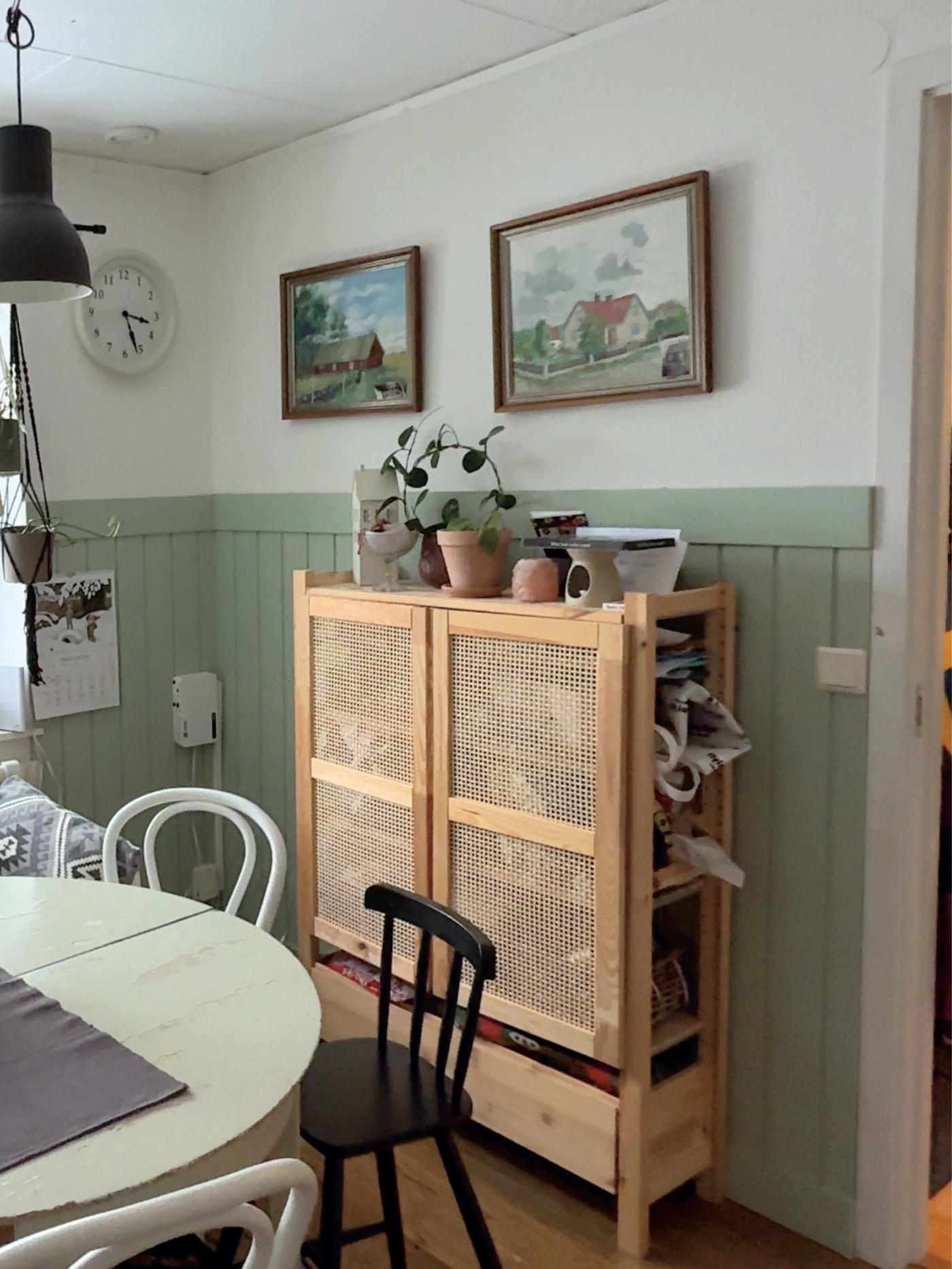 Hörna av matplats med rund bordskant i förgrunden, en ljus träförvaring med flätade dörrar och växter ovanpå samt två tavlor på väggen