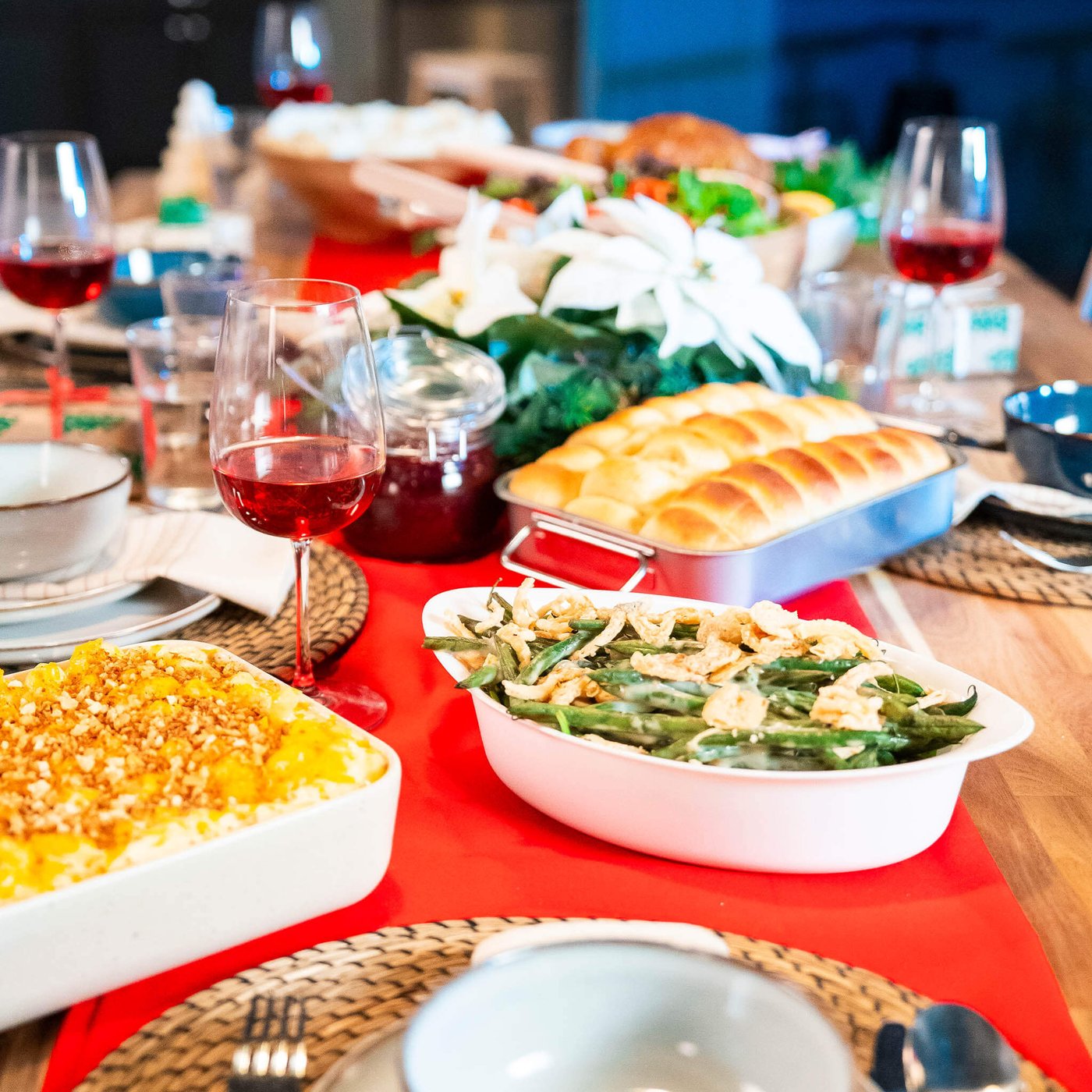 Holiday dining table with wine glasses, bread rolls, green bean casserole, mac and cheese dish, and festive centerpiece.