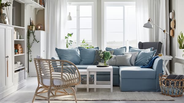 HOLMSUND light blue corner sofa-bed in a living room setting, with a rattan chair, white coffee tables and a large window.
