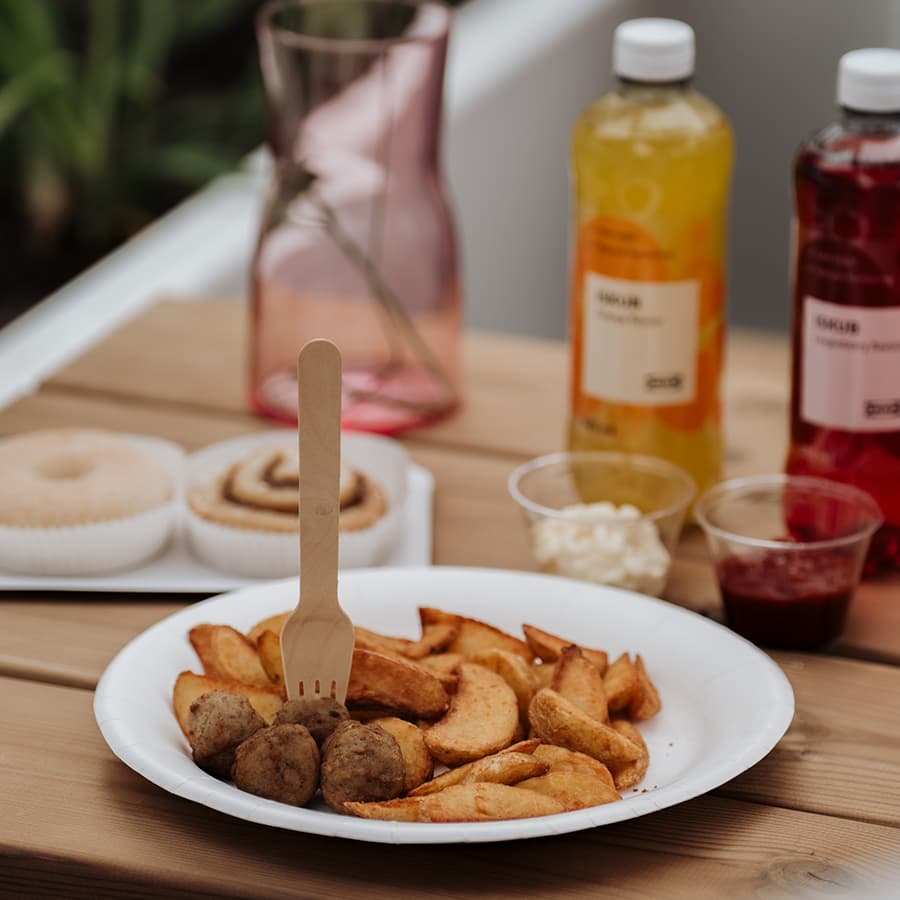 Holztisch mit einem weißen Teller, auf dem Bällchen und Wedges liegen. Daneben befinden sich Dip-Schalen mit Soßen, eine ISKUB Flasche, eine rosafarbene Vase sowie Süßwaren im Hintergrund.