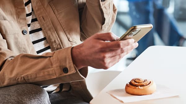 Hombre bebiendo un café caliente en el restaurant IKEA, con su celular en la mano y frente a el un rollito de canela IKEA sobre una servilleta.