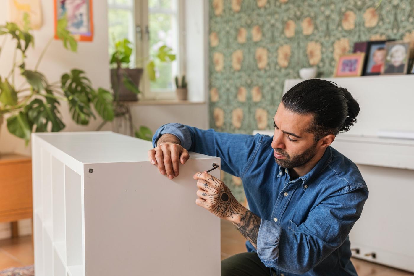 Hombre de piel morena y pelo negro con cola, vestido con una camisa de jean armando un mueble blanco.