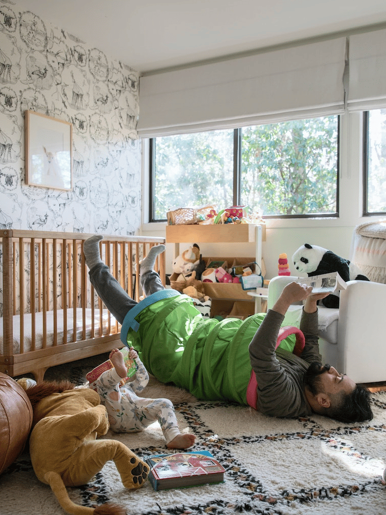 Hombre dentro de una cuncuna de juegos para niños recostado en el piso sobre una alfombra de color blanco con gris, a su lado un bebé en la misma posición que el y peluches.