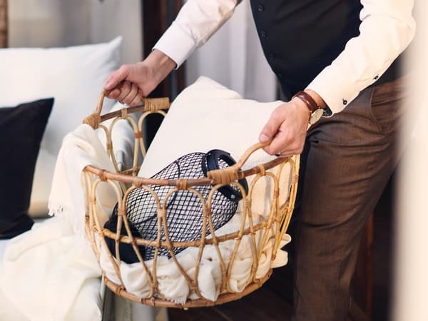 Hombre sosteniendo un canasto de mimbre con un cojín blanco y una pantalla de lampara en su interior.