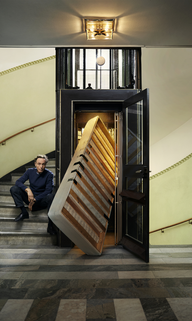 Home assegut en una escala al costat d’un ascensor antic on un somier amb matalàs queda encallat a la porta, en un ambient clàssic amb terra de marbre.