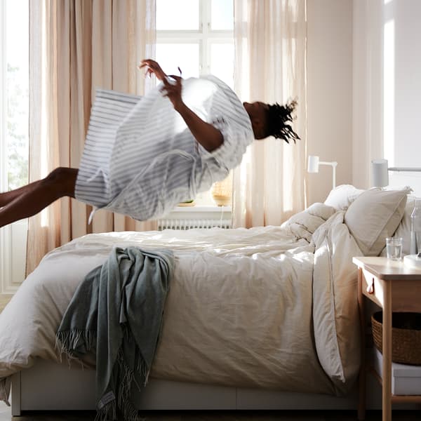 Homem a atirar-se de costas para cima de uma cama MALM com um colchão de molas ensacadas VATNESTRÖM e roupa de cama em branco.