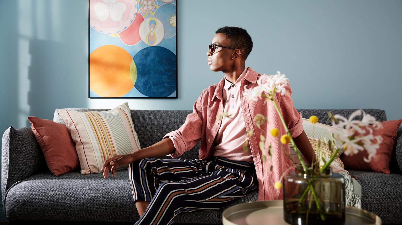 Homem com roupa em rosa e às riscas está sentado num sofá em cinzento, encostado a uma parede com um póster grande de arte a pastel.