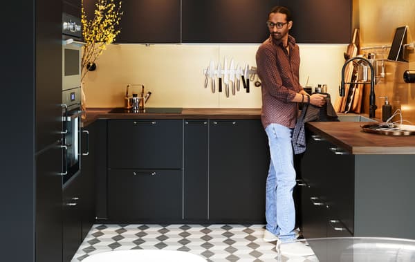 Homem de pé numa cozinha em forma de U, moderna e organizada, em cores escuras. Chão com mosaicos em xadrez, iluminação discreta.