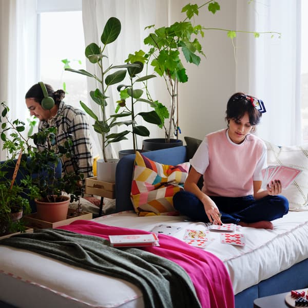 Homem e mulher sentados em duas camas dispostas costas com costas, com plantas entre eles. A mulher está de pernas cruzadas e a jogar às cartas.