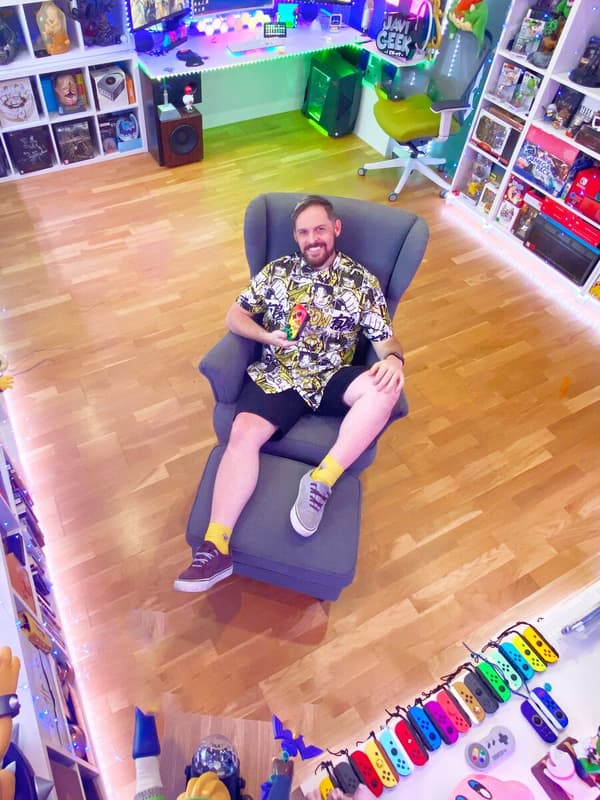 Homem sorridente recostado numa poltrona STRANDMON. Tem uma consola de videojogos na mão. As paredes à volta estão cobertas de prateleiras com videojogos e brinquedos.