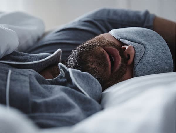 Homme enpyjama bleu avec un masque sur les yeux, en train de dormir la bouche ouverte
