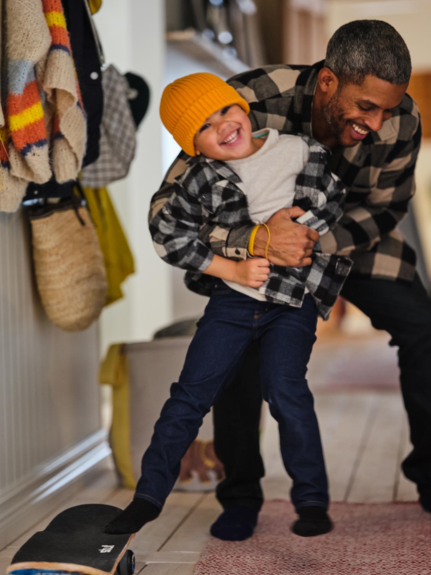 Homme et enfant jouant à la lutte dans un hall d’entrée. Autour d’eux, des vêtements suspendus, un skateboard et un sac de rangement JÄTTELIK à motif de dinosaures.