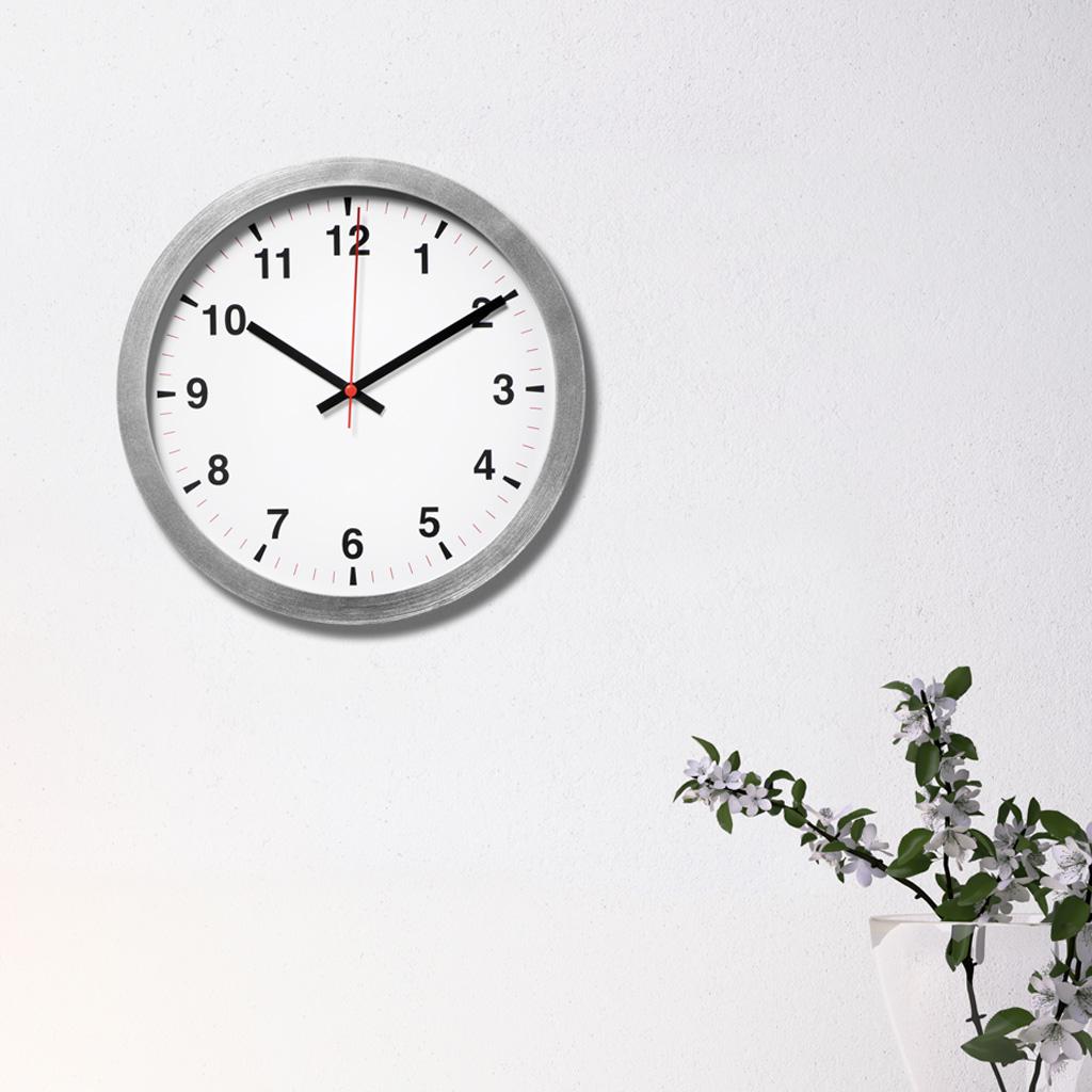 Hopeinen TJALLA-seinäkello valkoisella seinällä. Kello näyttää kymmentä yli kymmenen.