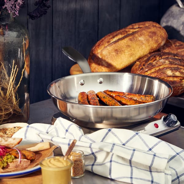 Hot dog vegetariani KORVMOJ in padella sopra un fornello a induzione. Accanto, pagnotte, salse e piatti con alimenti.