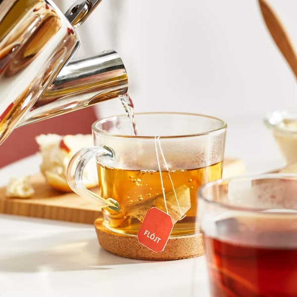 Hot water poured into glass with FLÖJT tea bag on cork coaster; red drink and food items on wooden board in background.