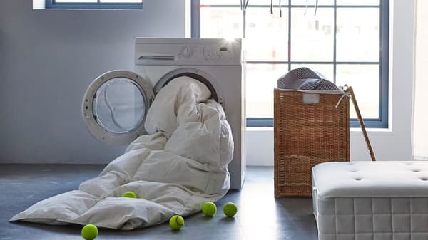 How to wash a duvet. An open dryer has a white duvet spilling out and tennis balls on the floor. A laundry hamper and a mattress are placed close to the dryer.