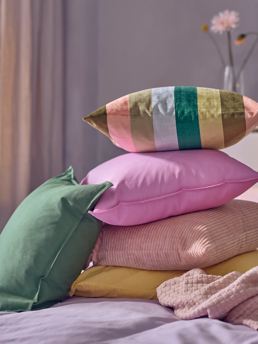 Hromádka polštářů v různých barvách a vzorech na posteli. Je vidět zelený, sytě růžový, žlutý, ale i s vícebarevnými pruhy a světle růžový s manšestrovým povrchem.