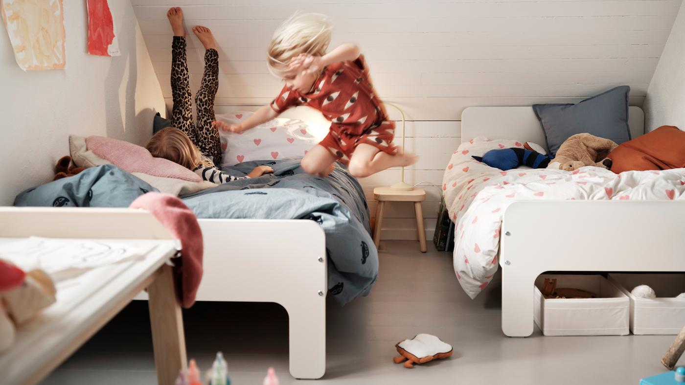Huoneessa, jossa on kaksi jatkettavaa SLÄKT-sänkyä, lapsi makaa jalat seinää vasten, kun toinen lapsi hyppää samalle sängylle.