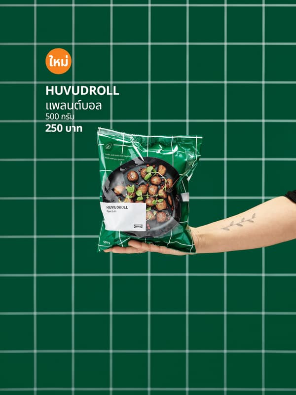 มือที่ยืนมา ในมือถือห่อบอลผัก HUVUDROLL ที่ยังไม่ได้เปิด พื้นหลังเป็นผนังกระเบื้องสีเขียว