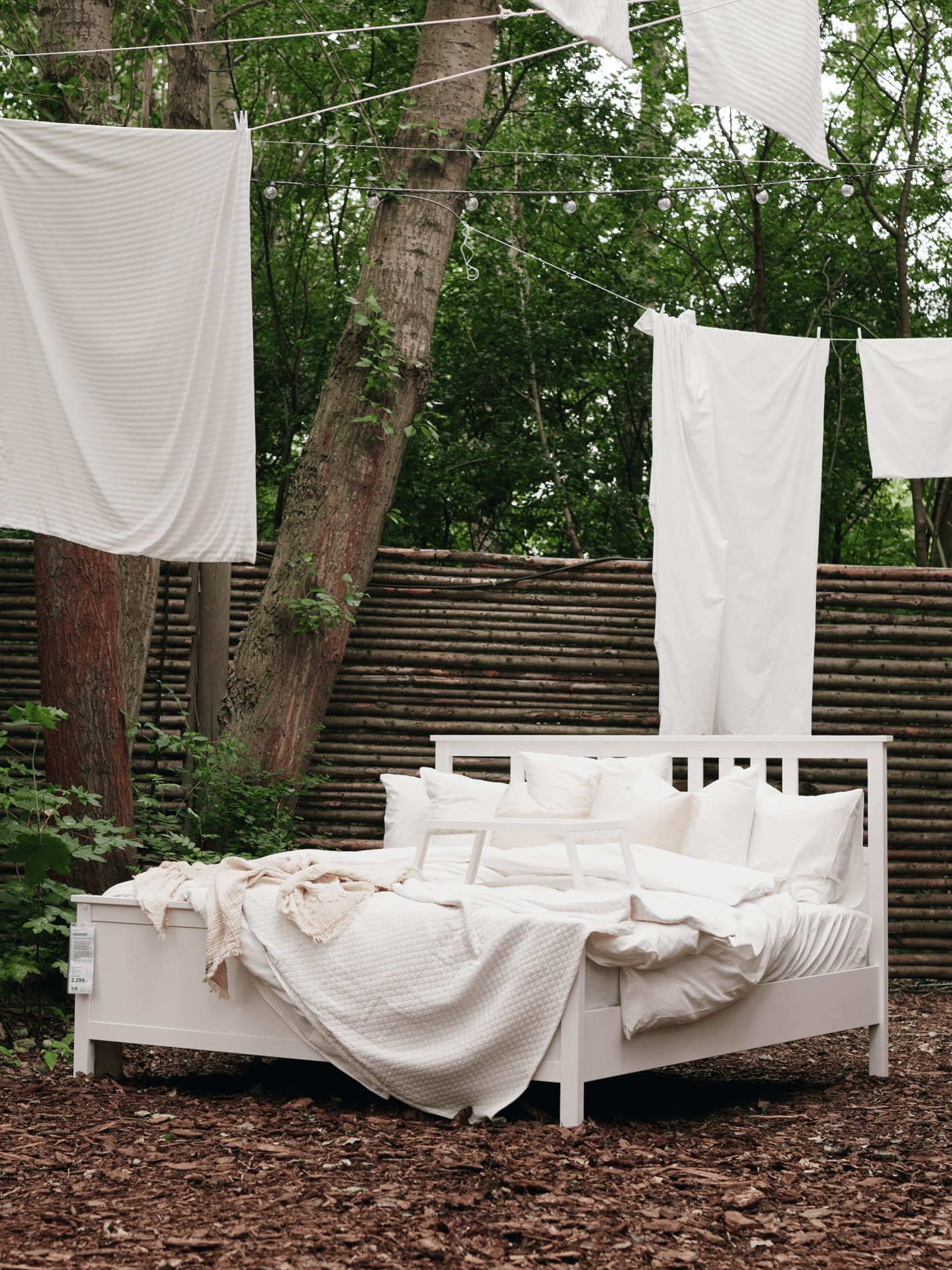 I et udendørs miljø står en seng med hvidt sengetøj dyner og puder over sengen hængerforskellige hvide dyner.