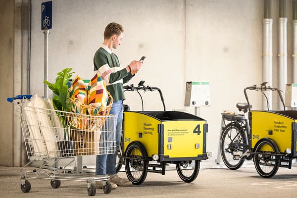I ett parkeringsgarage står en person med en telefon i handen. Bredvid personen står en kundvagn och två lastcyklar.