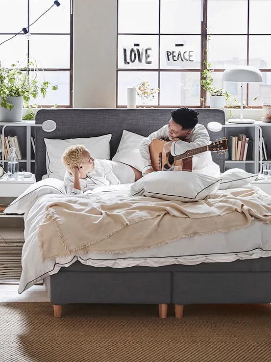 I ett rum med stora fönster ligger två personer och tittar på varandra i en säng. Den ena personen håller om en gitarr.