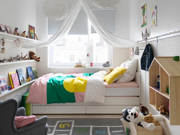 白いベッドにピンクとグリーンの寝具を合わせた子ども部屋。窓辺に本棚とぬいぐるみを配置。