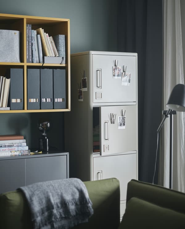 IDÅSEN beige high cabinet with smart locks stands by a wall. Magnetic clips with photos are put on some of the cabinet doors.