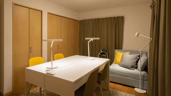 白いデスクが2セットと黄色の椅子が2脚置かれている。奥にグレーのソファが置かれ、クッションが並んでいる。