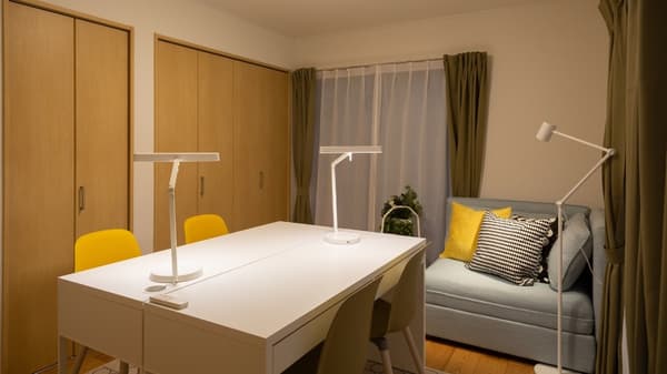 白いデスクが2セットと黄色の椅子が2脚置かれている。奥にグレーのソファが置かれ、クッションが並んでいる。