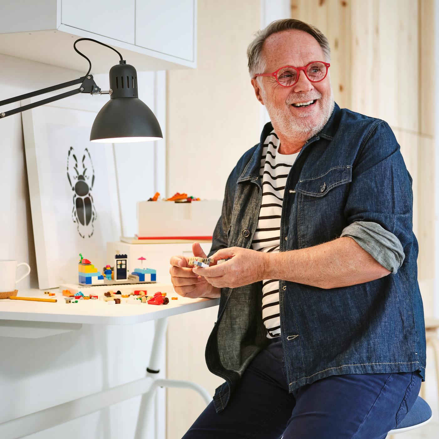 Iemand in een jeanshemd en -broek zit op een NILSERIK zit-stasteun en speelt met BYGGLEK LEGO steentjes aan een wit GLADHÖJDEN bureau.