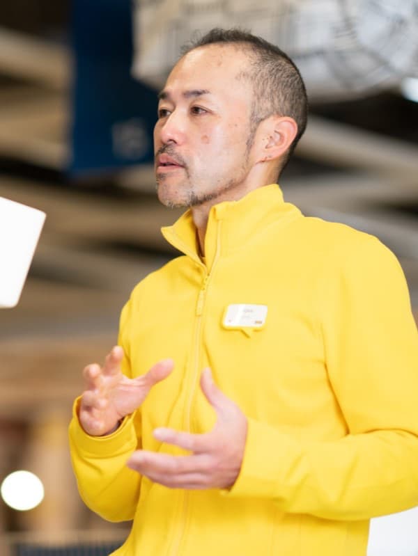 イエローのジャケットを着たコワーカーが、イケアストアのフロアで名札を付けて説明を行い、周囲にホームファニッシング商品のディスプレイが見える。
