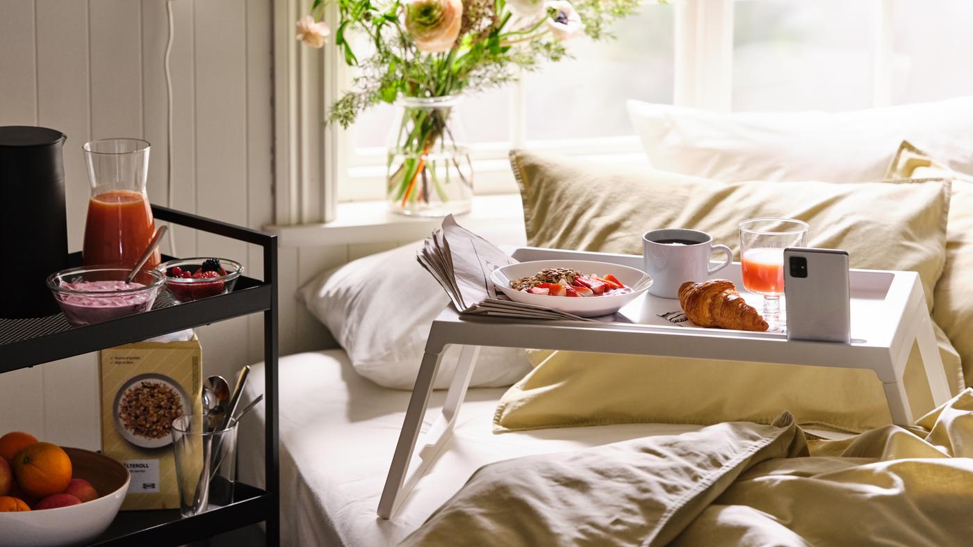إفطار يُقدم على صينية سرير KLIPSK بيضاء على سرير مرتب عليه مفارش سرير ÄNGSLILJA، مع المزيد من الطعام على عربة.