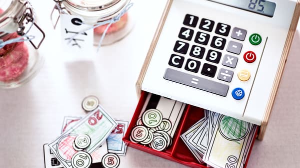 Igračka blagajna s velikim tipkama i zaslonom koji prikazuje broj 85, otvorena ladica s papirnatim novčanicama i okruglim kovanicama, te staklene tegle s etiketama na stolu.