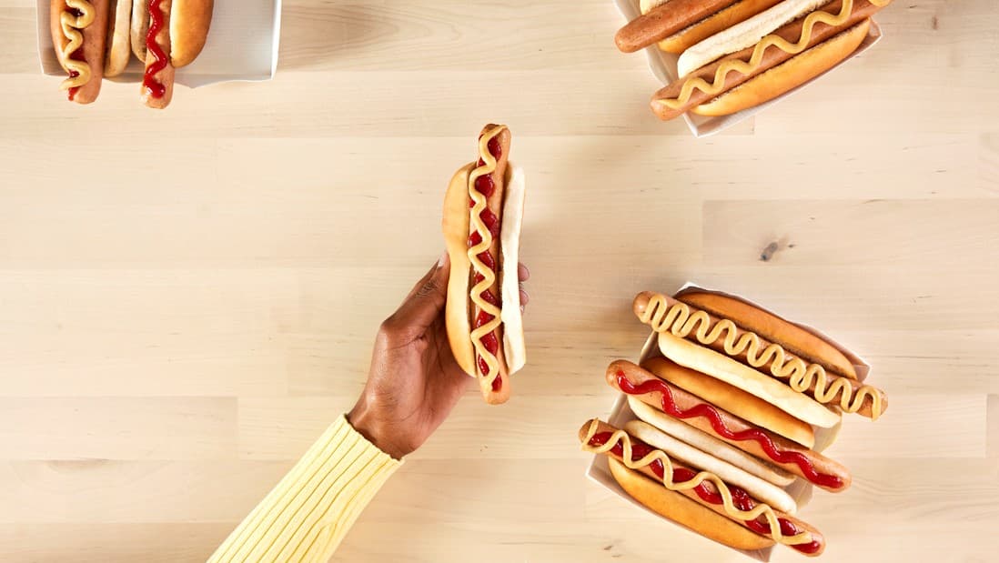 Ihminen pitää hot dogia kädessään pöydän päällä.