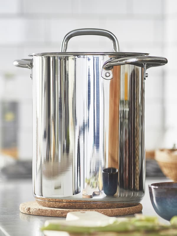 סיר IKEA 365+ עם מכסה עומד על מעמד סירים HEAT המונח על ספסל מטבח אפור ליד קערה עם זיגוג כחול כהה.