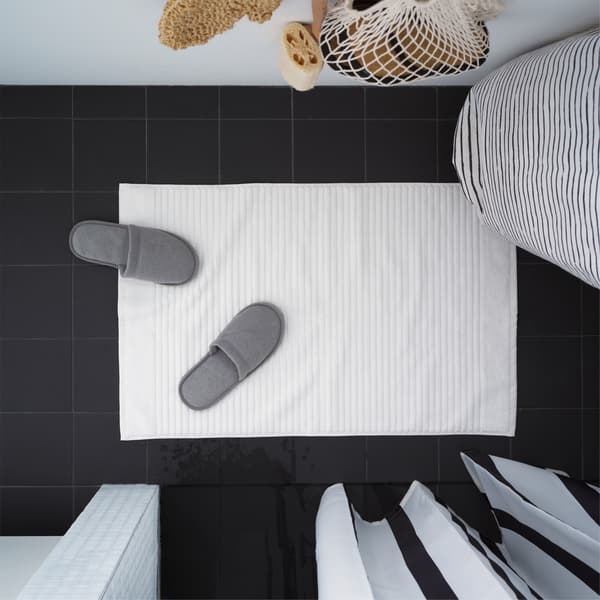 IKEA ALSTERN white bath mat is placed on a black bathroom floor. Grey slippers are placed on the mat.