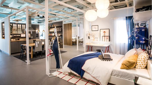 IKEA-Ausstellungsbereich mit vollständig eingerichteter Wohnsituation. Im Vordergrund ein Bett mit Deko-Kissen, im Hintergrund Küche, Essplatz und Arbeitsbereich. Helle Hängelampen sorgen für gemütliche Atmosphäre.