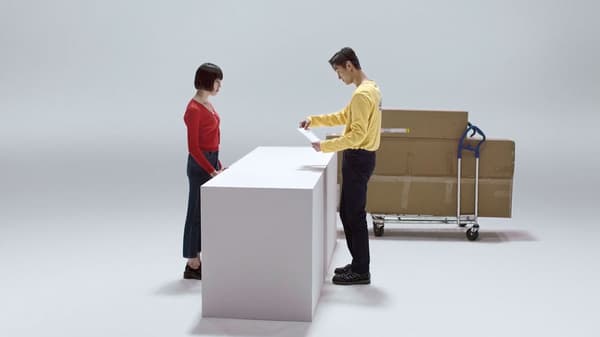 IKEA co-worker in a yellow shirt assisting a customer at a counter, with flat-packed furniture boxes on a trolley in the background.
