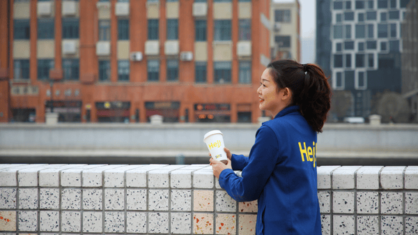 IKEA co‑worker in blue jacket holding a “Hej!” coffee cup, relaxing by a riverside wall with an urban cityscape in the background.